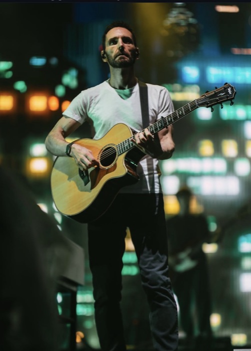 Johnny McDaid standing with an acoustic guitar against city lights