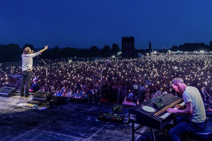 Johnny McDaid at the keys on a festival stage at dusk, before a crowd holding up phone lights